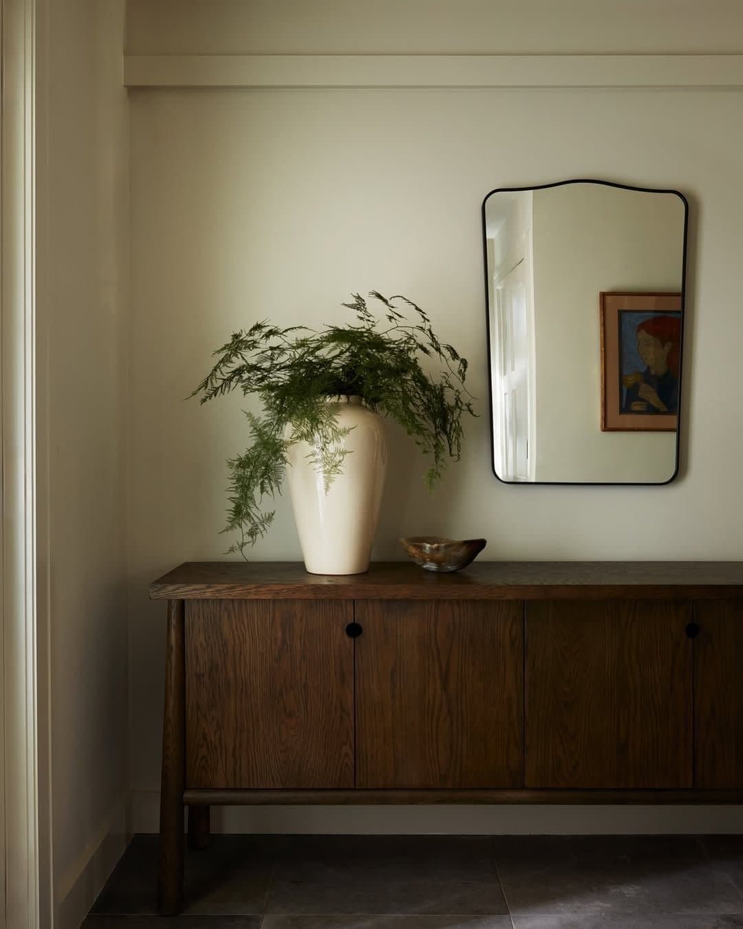 A warm wood console styled with a mirror, vessel, and greenery